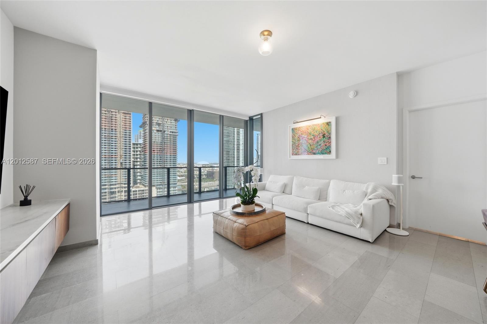 A modern living room features white walls, tile flooring, a white sofa, and floor-to-ceiling windows — 68 SE 6th St # 1904, Miami FL 33131, MIAMI, FL