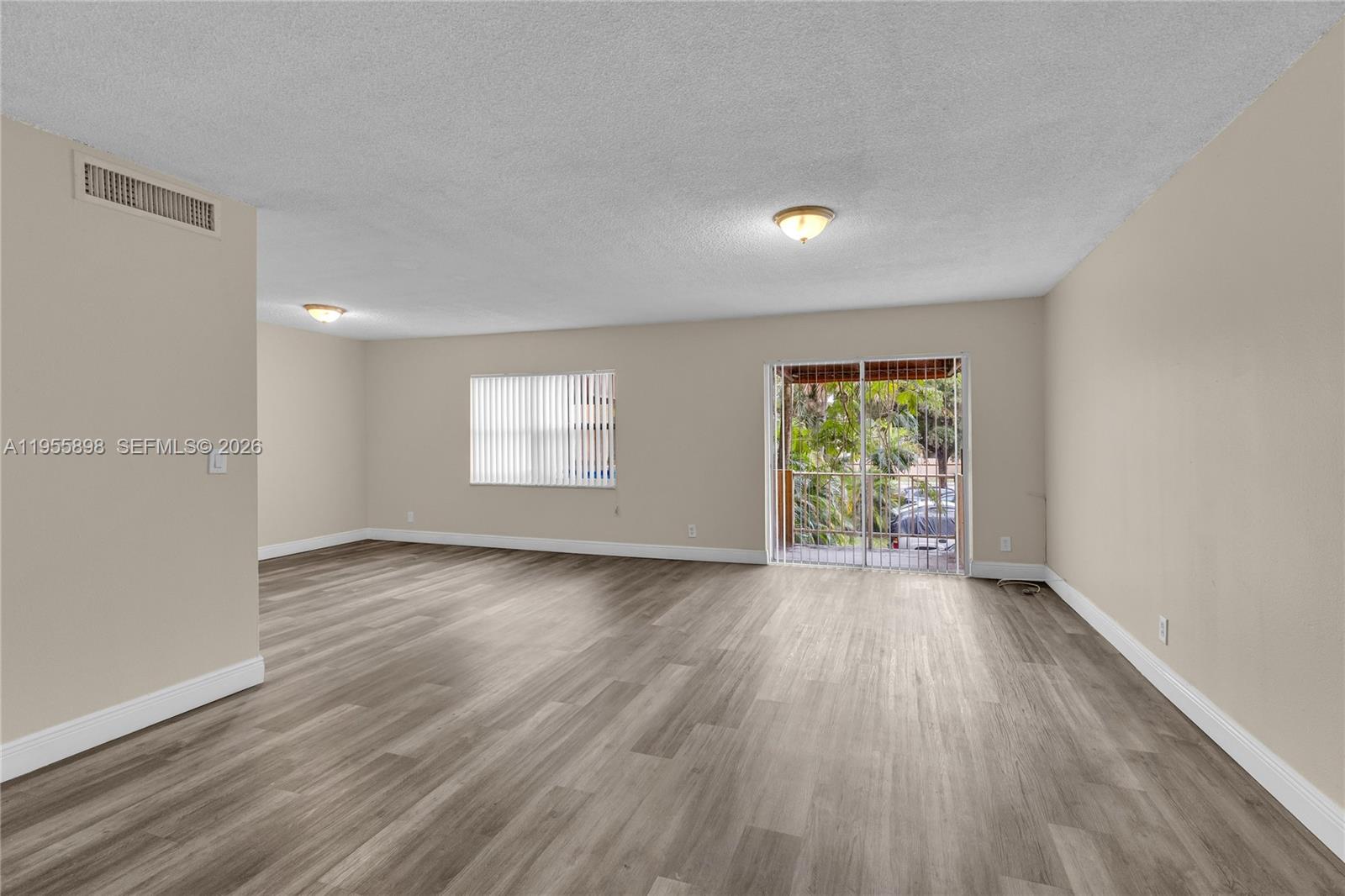 An empty living room features light-colored walls, wood-look flooring, a window, and a sliding glass door — 17901 NW 68th Ave # T206, Hialeah FL 33015, HIALEAH, FL