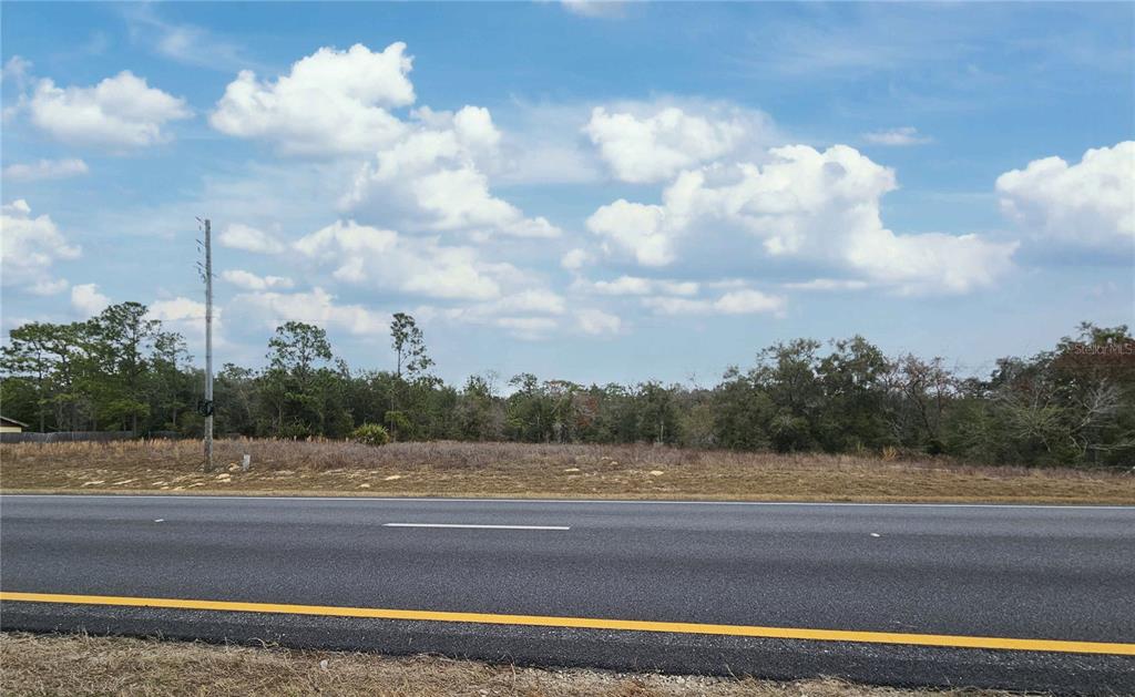 A vacant, grassy lot sits behind a paved road under a blue sky with clouds — 0000 NE HIGHWAY 27 ALT, WILLISTON, FL