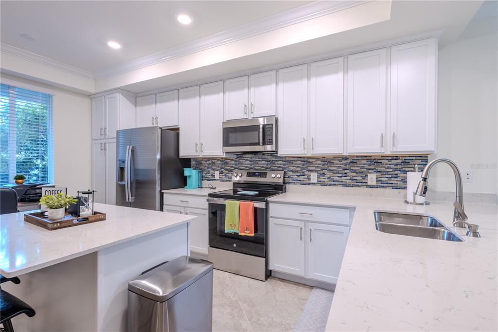 A modern kitchen features white cabinets, stainless steel appliances, quartz countertops, and a blue backsplash — 1030 TIDEWATER SHORES LOOP UNIT 104, BRADENTON, FL
