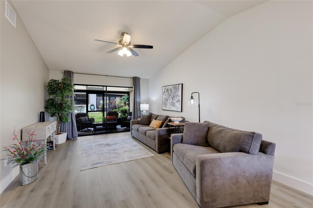 A living room features gray sofas, wood-look flooring, a ceiling fan, and a sliding glass door — 6427 HERITAGE LANE UNIT 6427, BRADENTON, FL