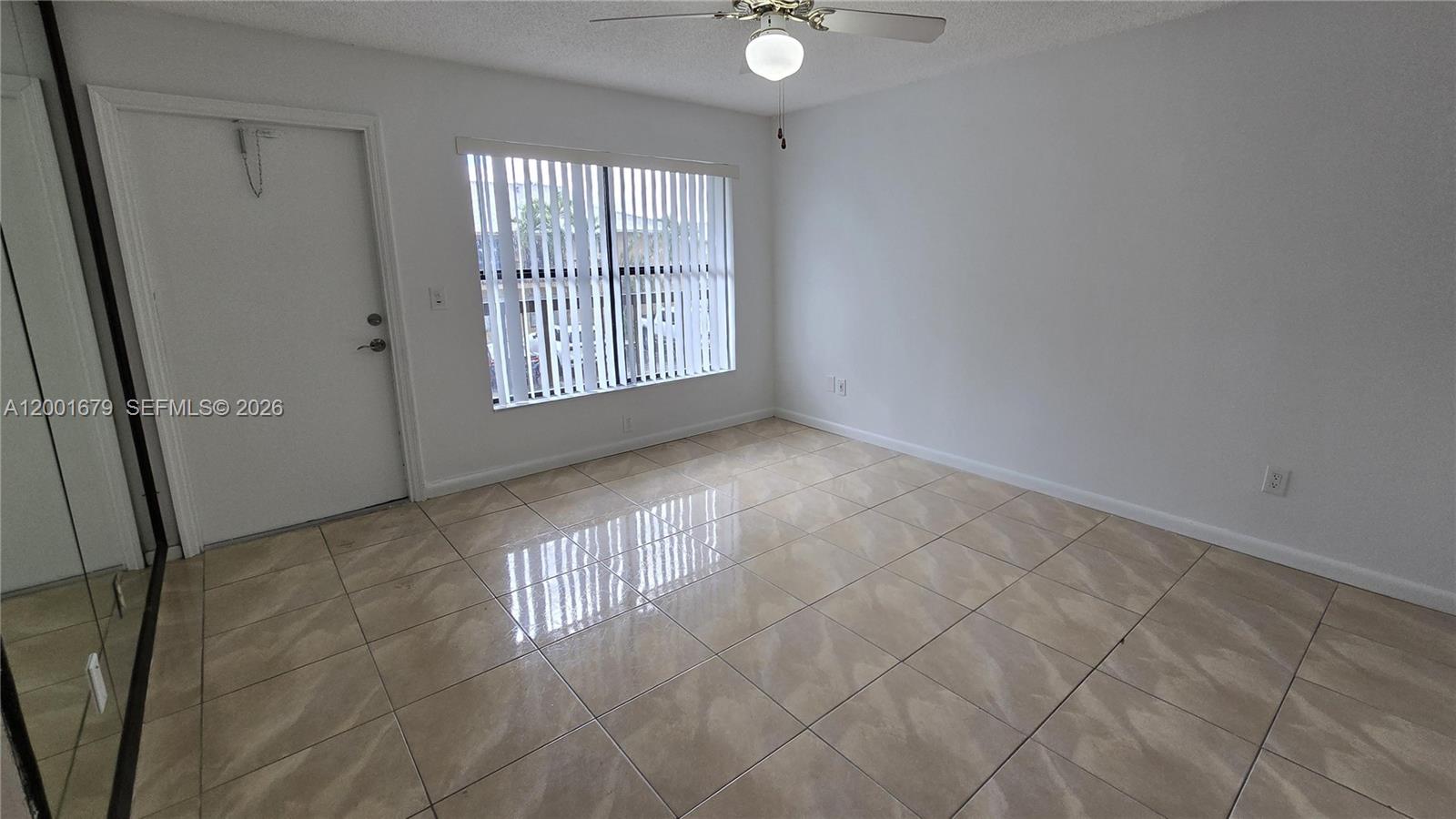 An empty room features white walls, beige tile flooring, a ceiling fan, and vertical blinds — 2605 W 67th Pl # 10, Hialeah FL 33016, HIALEAH, FL