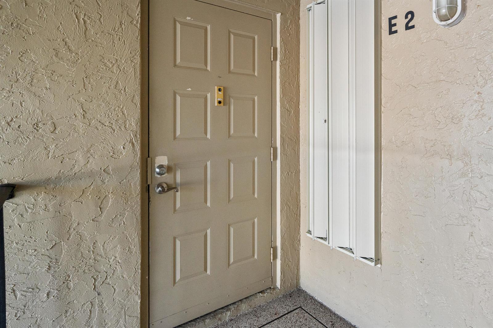 A beige door with a peephole and metal hardware is labeled E2 on a textured wall — 706 Sunny Pine Way # E2, FL 33415, FL