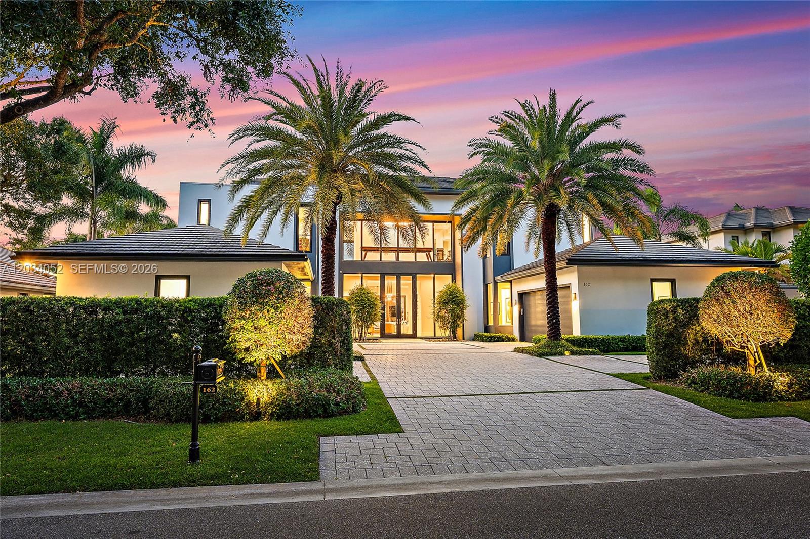 A modern two-story house with a paved driveway, palm trees, and large glass windows at dusk — 162 Commodore Dr, Jupiter FL 33477, JUPITER, FL