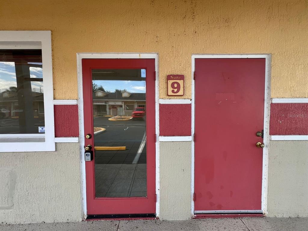 Two red doors are set into a tan stucco wall with a suite number sign — 4400 N HIGHWAY 19A UNIT 9, MOUNT DORA, FL