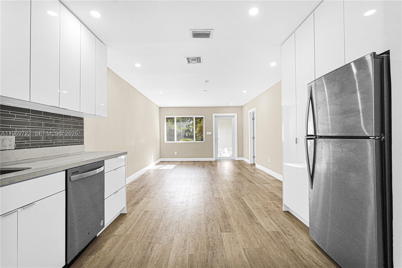 A modern kitchen with white cabinets, stainless steel appliances, and wood-look flooring opens to a living area — 11919 NE 8th Ave, Biscayne Park FL 33161, BISCAYNE PARK, FL