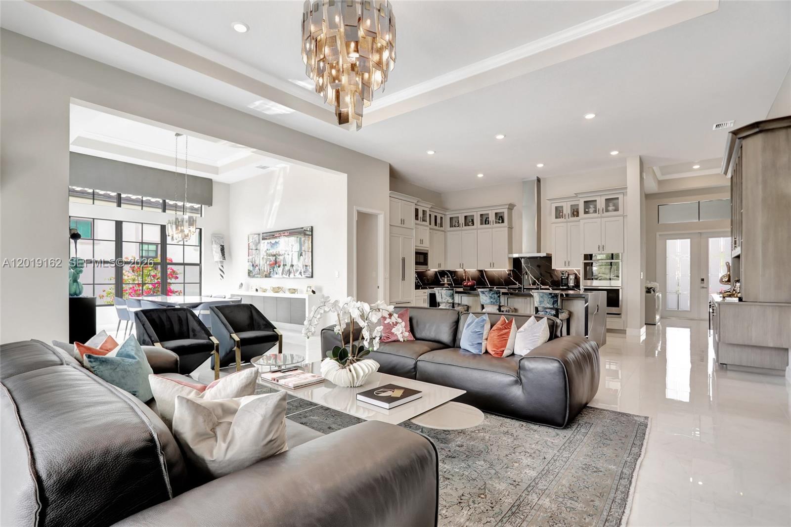 A modern open-concept living room features a dark leather sofa, a chandelier, and a kitchen — 8044 Ironwood Way, Parkland FL 33067, PARKLAND, FL