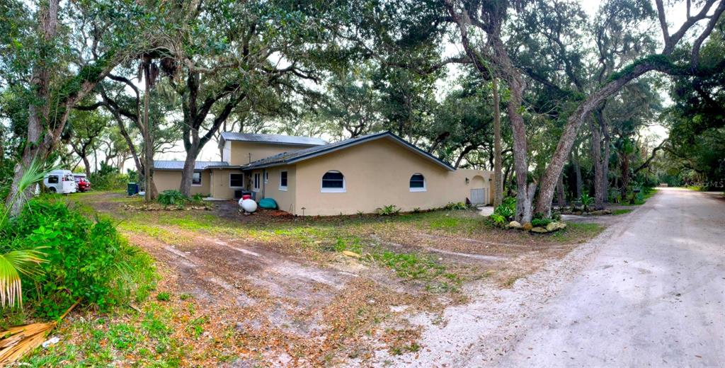A tan, single-story house with a metal roof sits among large trees near a dirt road — 3 ELIZABETH DRIVE E, PALM COAST, FL
