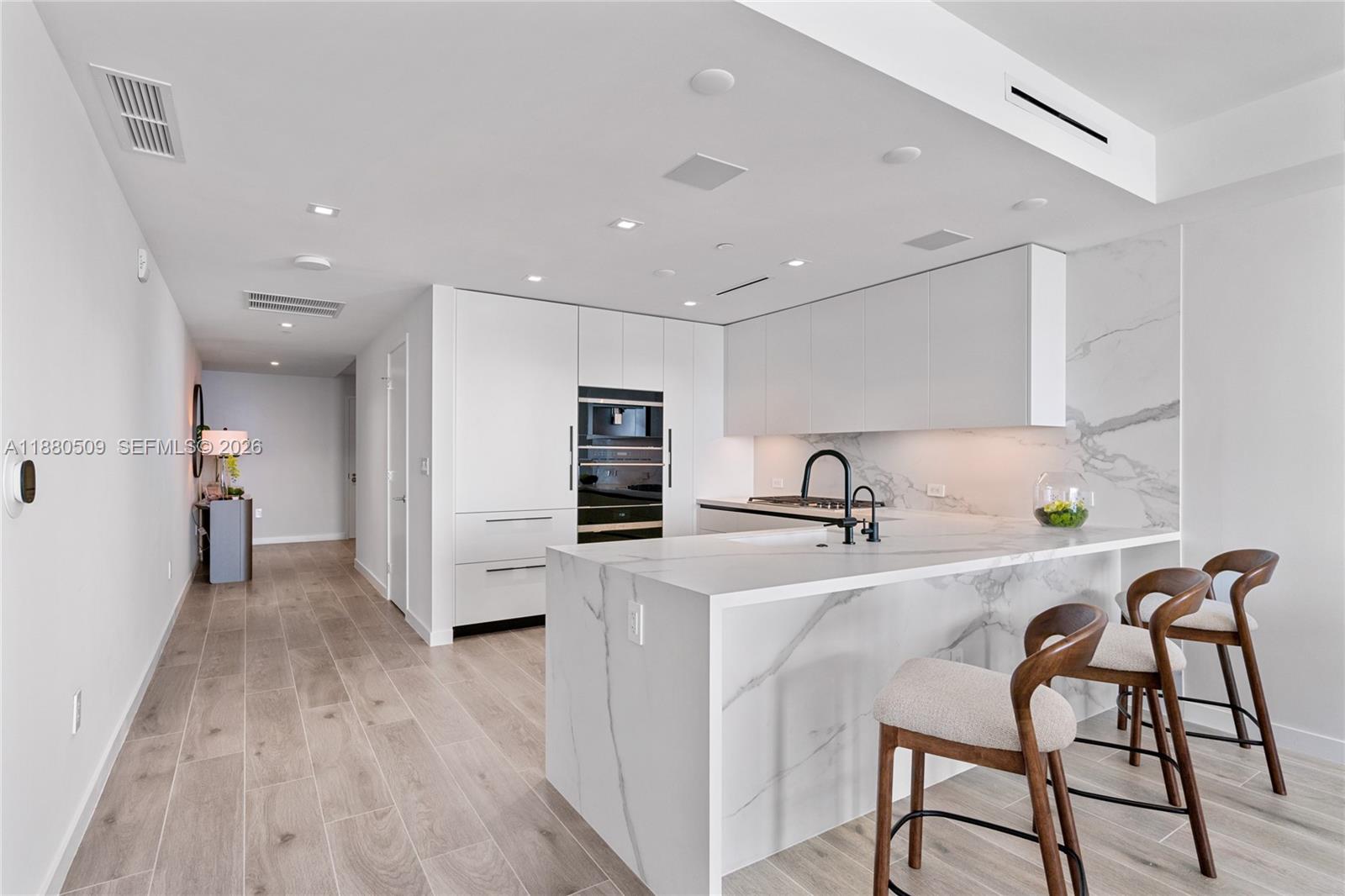 A modern white kitchen features a marble waterfall island, integrated appliances, and wooden bar stools — 1116 N Ocean Blvd # 1404, Pompano Beach FL 33062, POMPANO BEACH, FL