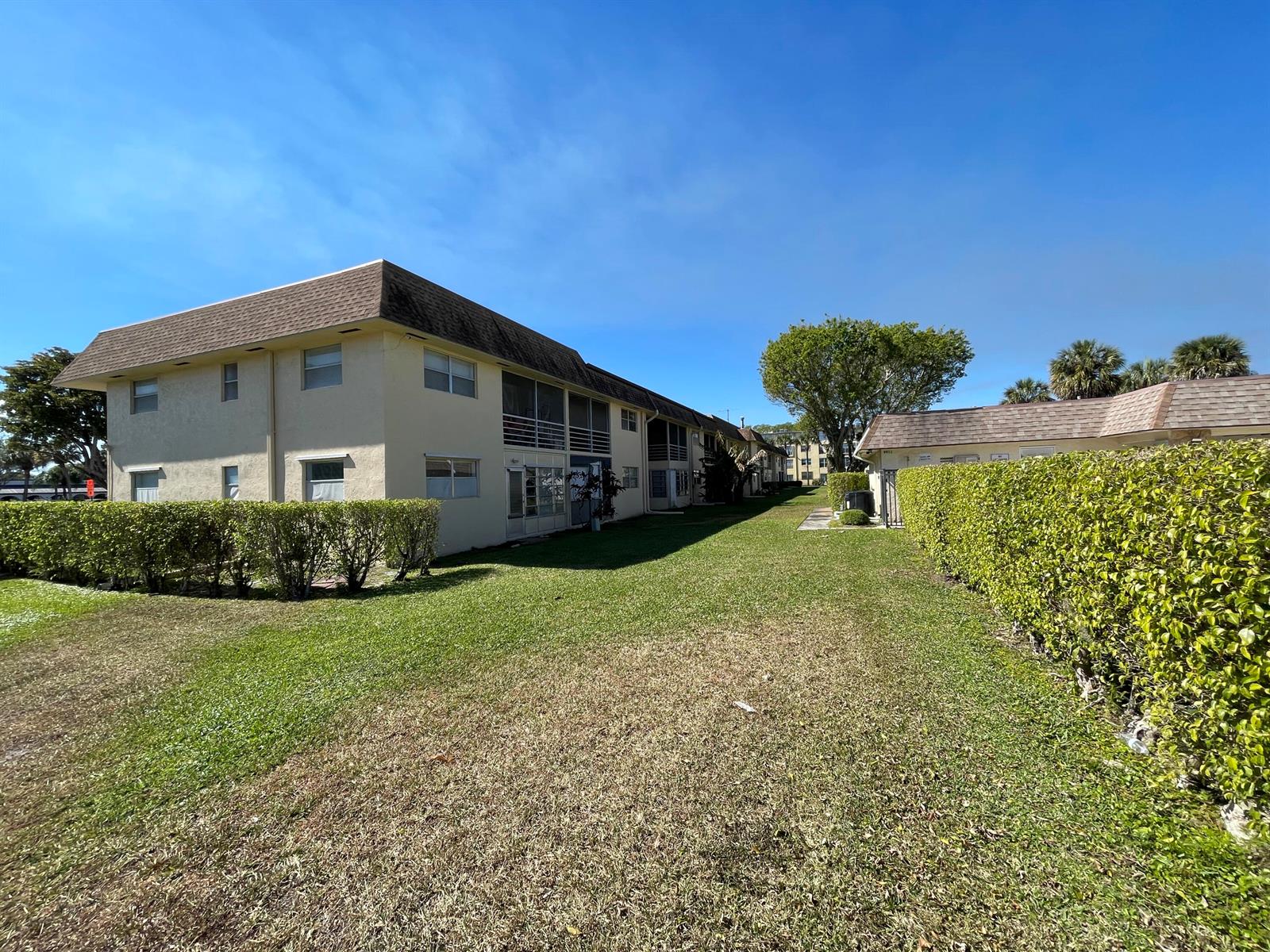A two-story apartment building sits beside a grassy lawn bordered by tall green hedges — 9911 Sandalfoot Boulevard # 112, Boca Raton FL 33428, BOCA RATON, FL
