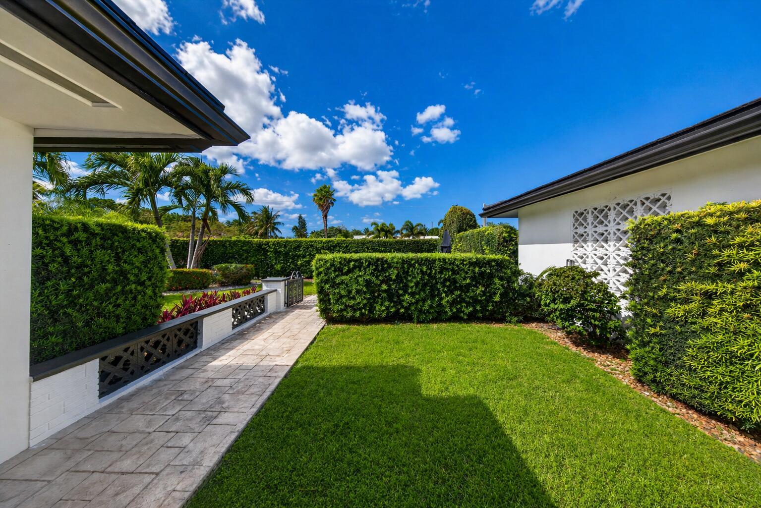 A paved walkway leads through a grassy backyard bordered by manicured hedges and white walls — 850 W Camino Real, Boca Raton FL 33486, BOCA RATON, FL