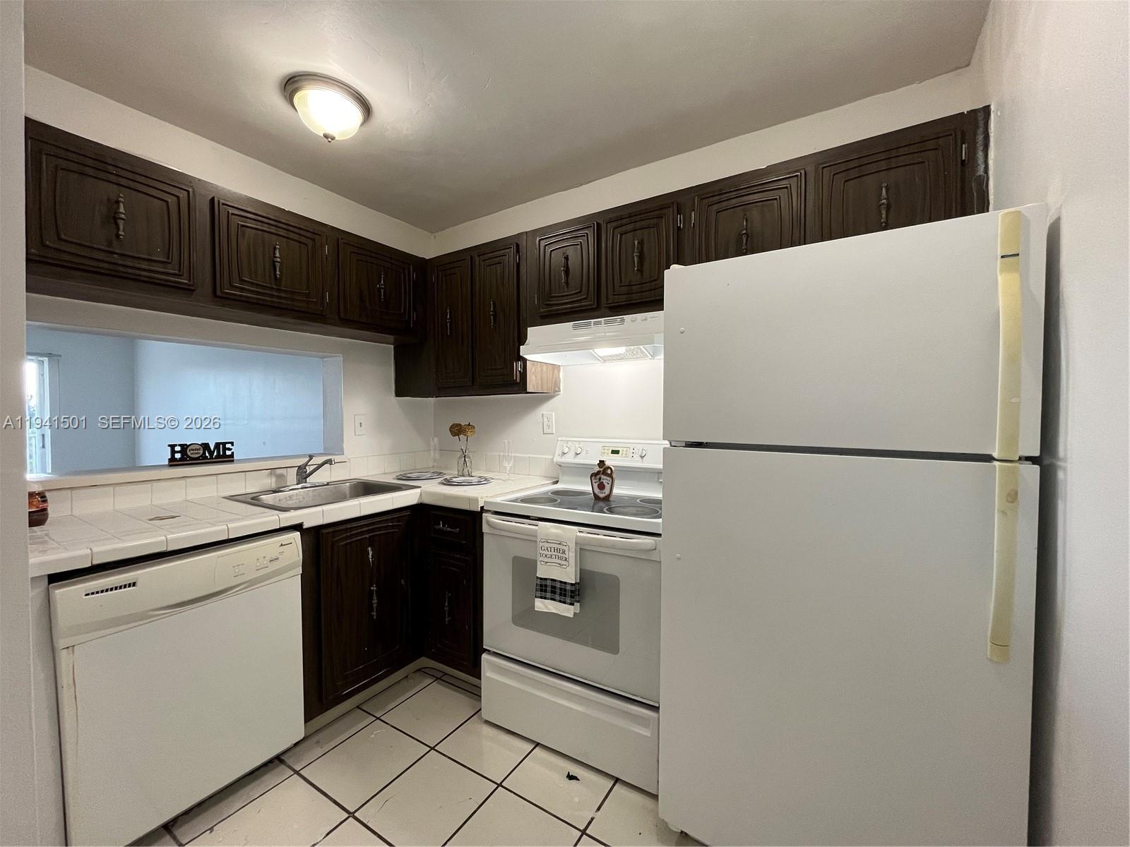 A kitchen features dark wood cabinets, white appliances, and white tiled countertops and flooring — 492 NW 165th St Rd # 611, Miami FL 33169, MIAMI, FL
