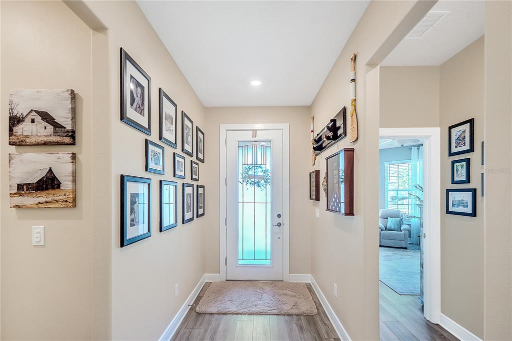 A bright entryway features wood-look flooring, a white front door, and walls decorated with framed photos — 9381 SW 56TH LOOP, OCALA, FL
