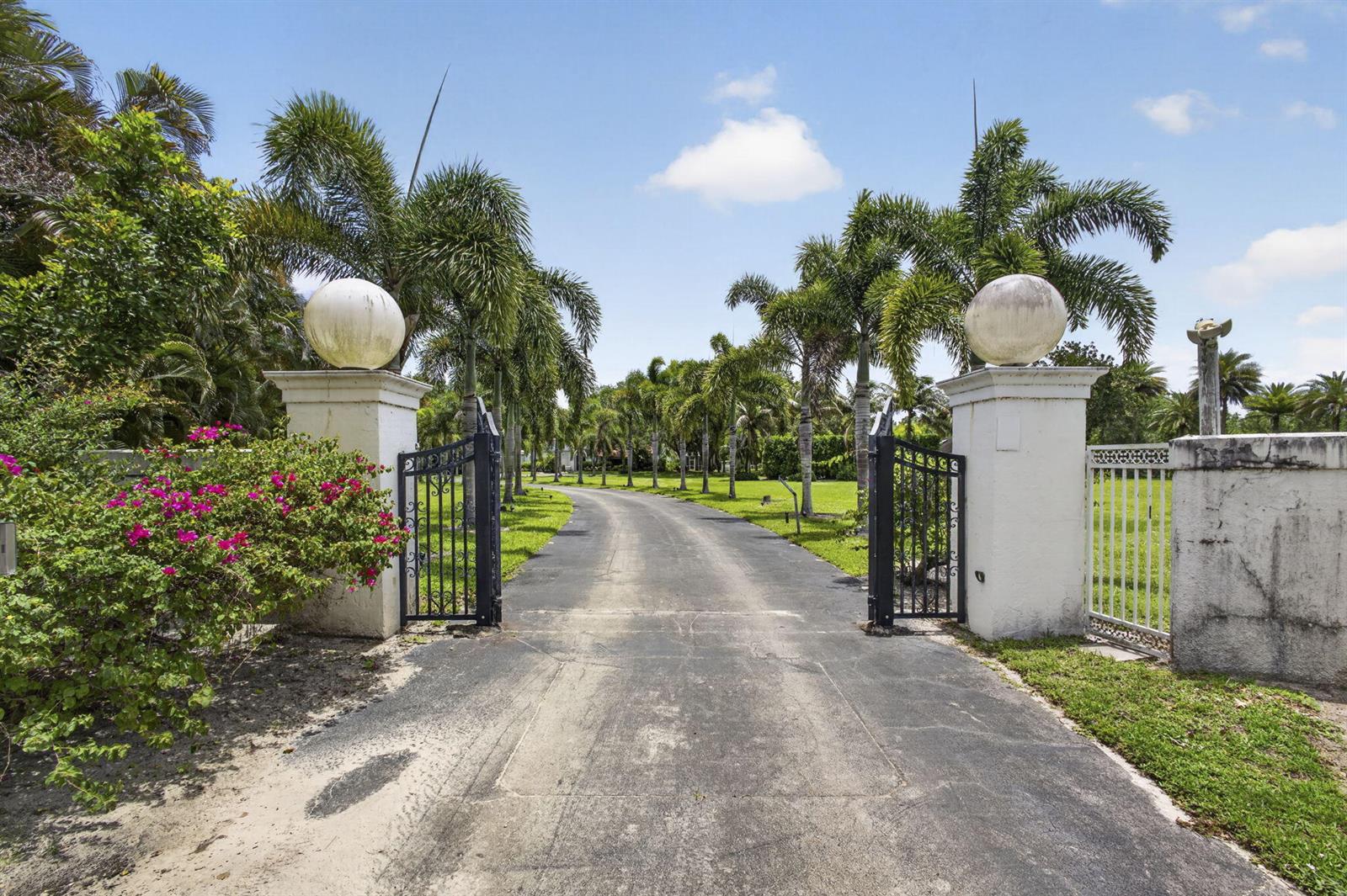 An asphalt driveway leads through an open gate flanked by white pillars with spherical finials — 1030 Arabian Drive, Loxahatchee FL 33470, LOXAHATCHEE, FL