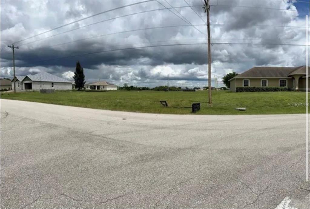 A vacant, grassy corner lot sits between two suburban houses under a cloudy sky — 2727 NW JUANITA PLACE, CAPE CORAL, FL