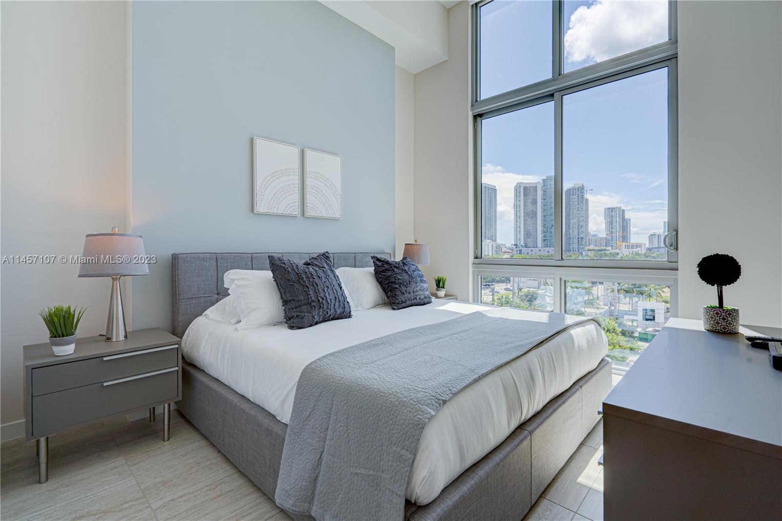 A modern bedroom features a gray upholstered bed, light blue wall, and floor-to-ceiling city views — 3900 Biscayne Blvd # 502, Miami FL 33137, MIAMI, FL