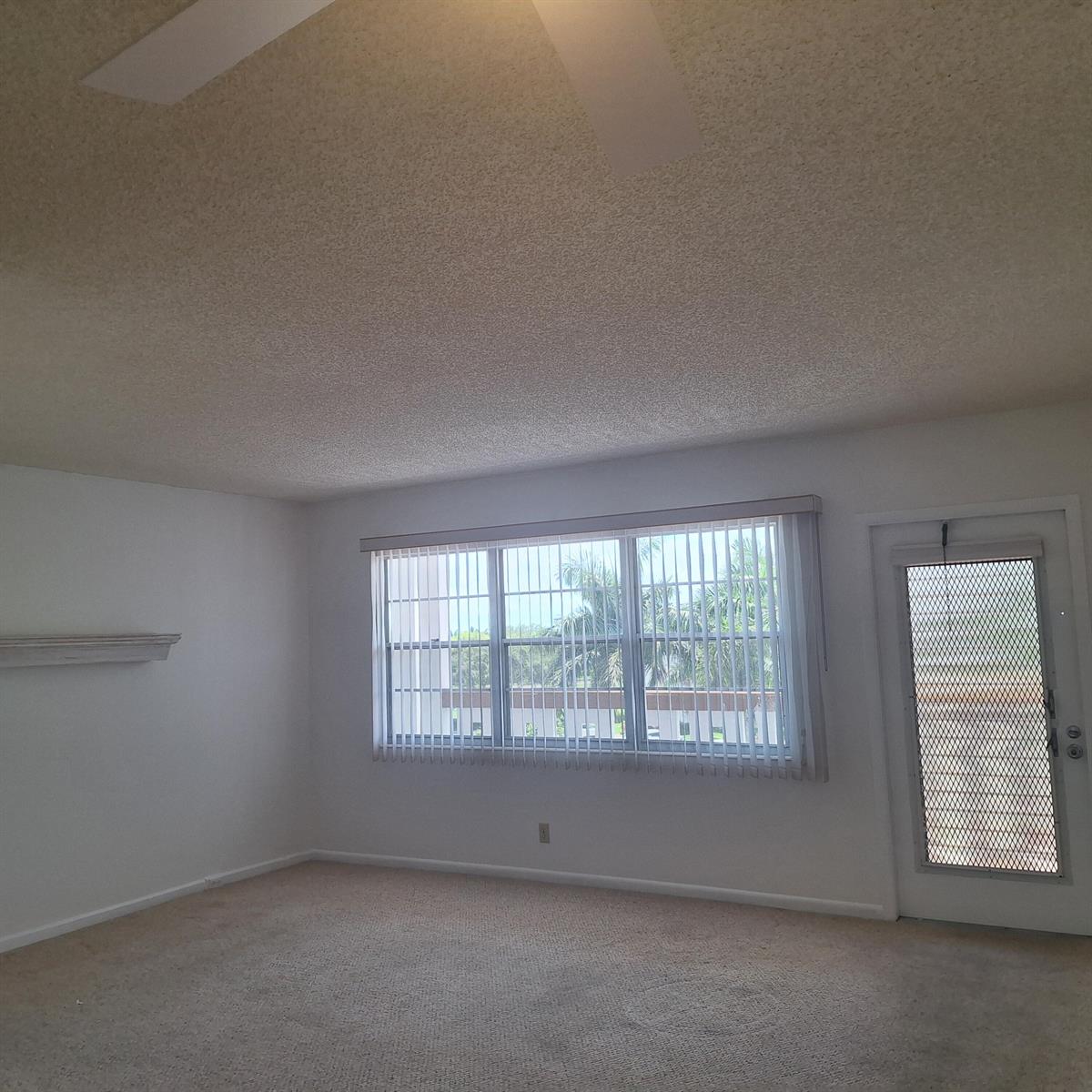 An empty room with beige carpet, white walls, a large window with blinds, and a door — 4007 Cornwall A # 4007, Boca Raton FL 33434, BOCA RATON, FL