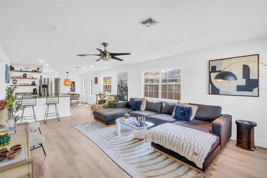A bright, open-concept living room features a large gray sectional sofa, light wood flooring, and kitchen — 218 49TH AVENUE N, ST PETERSBURG, FL