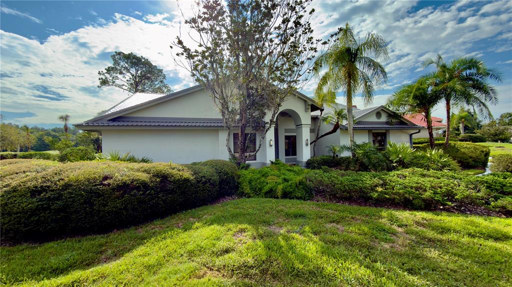 A single-story house with a metal roof, arched entryway, and extensive landscaping under a blue sky — 5040 PINELAKE ROAD, WESLEY CHAPEL, FL