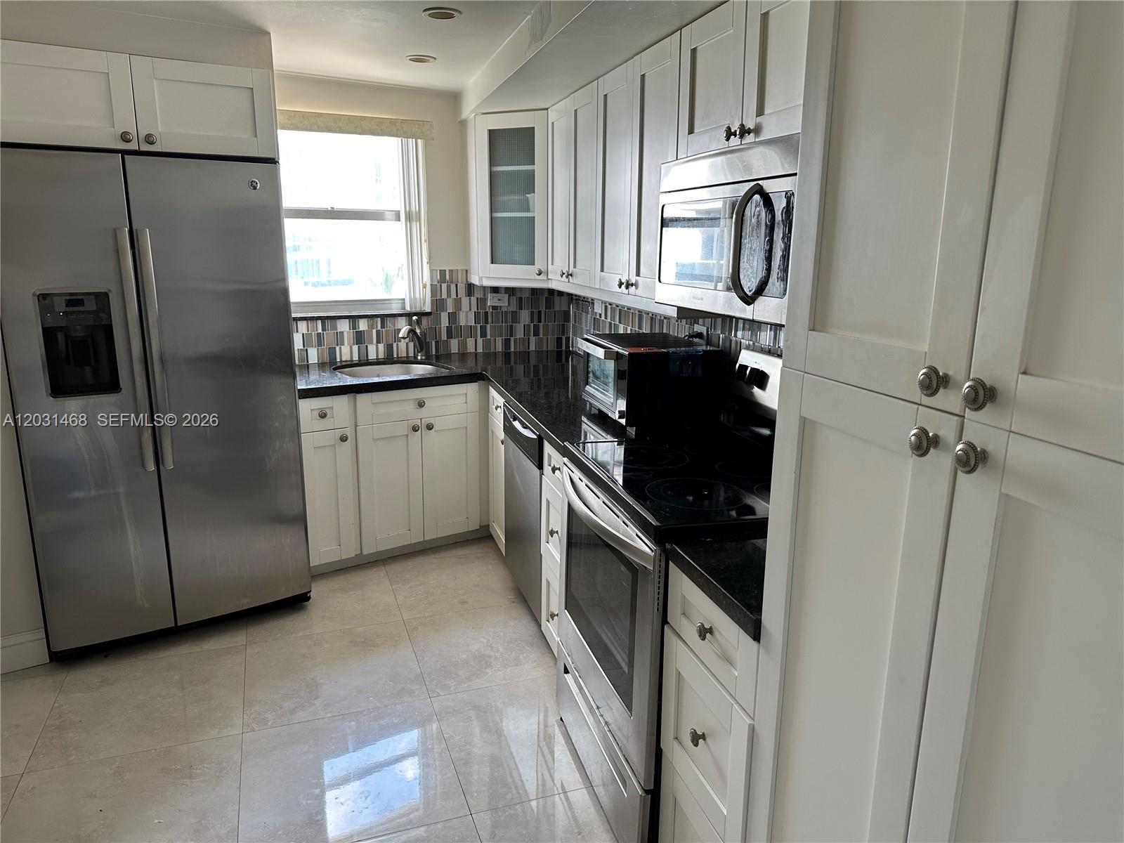 A kitchen features white cabinets, stainless steel appliances, black countertops, and light-colored tile flooring — 6039 Collins Ave # 728, Miami Beach FL 33140, MIAMI BEACH, FL