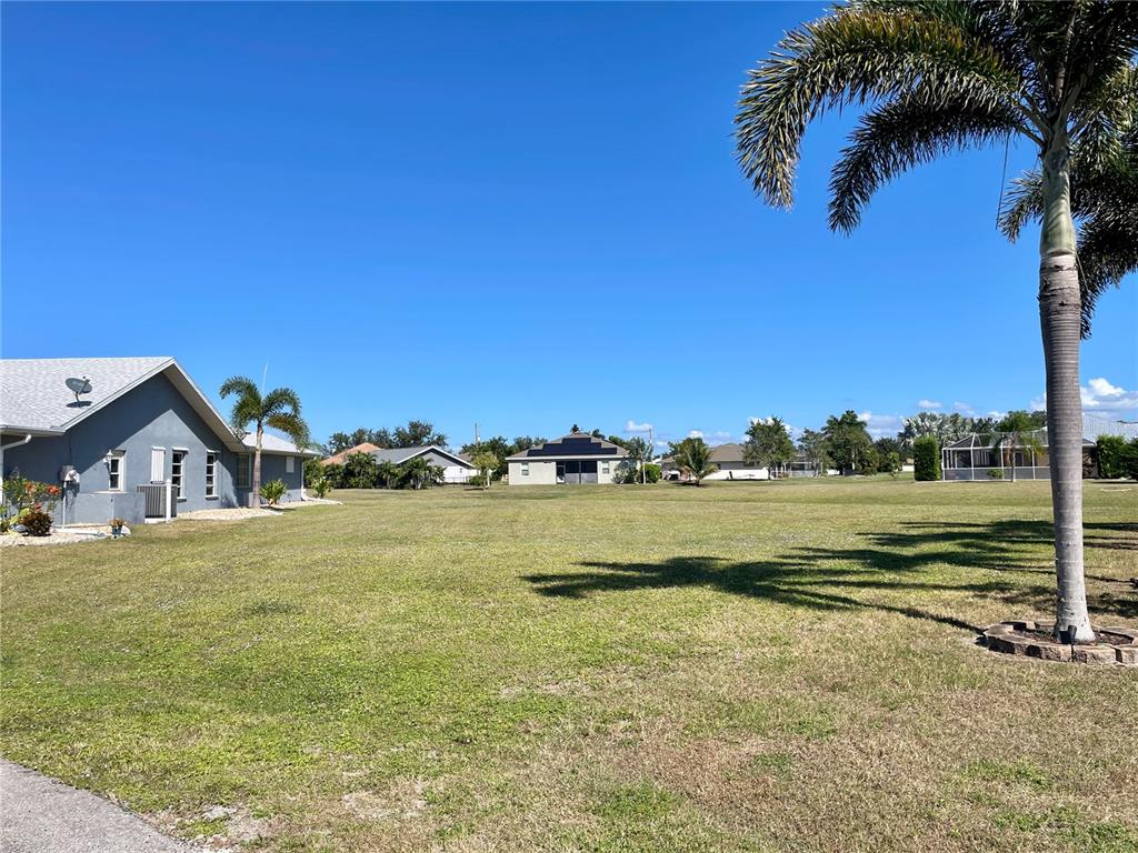 A large, grassy vacant lot sits between suburban houses under a clear blue sky — 7310 SATSUMA DRIVE, PUNTA GORDA, FL