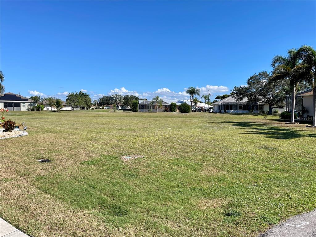 A wide, grassy vacant lot is surrounded by residential houses under a clear blue sky — 7310 SATSUMA DRIVE, PUNTA GORDA, FL