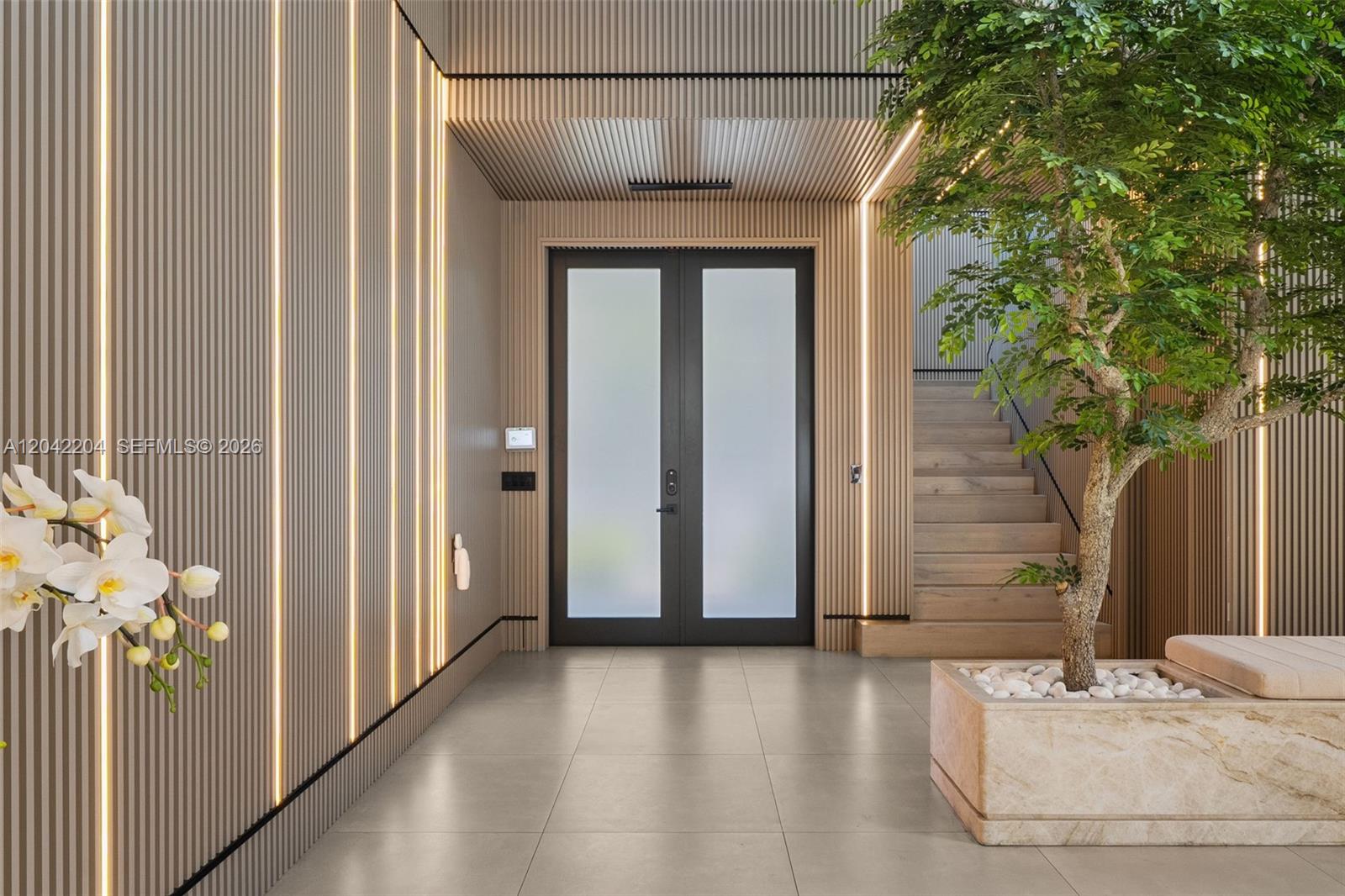 This modern entryway features slatted wood walls with integrated lighting, a tree, and stairs — 6814 NW 104th Ct, Doral FL 33178, DORAL, FL