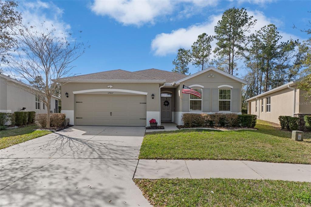 A single-story suburban house with a two-car garage, beige stucco exterior, and a front lawn — 17807 GARSALASO CIRCLE, BROOKSVILLE, FL