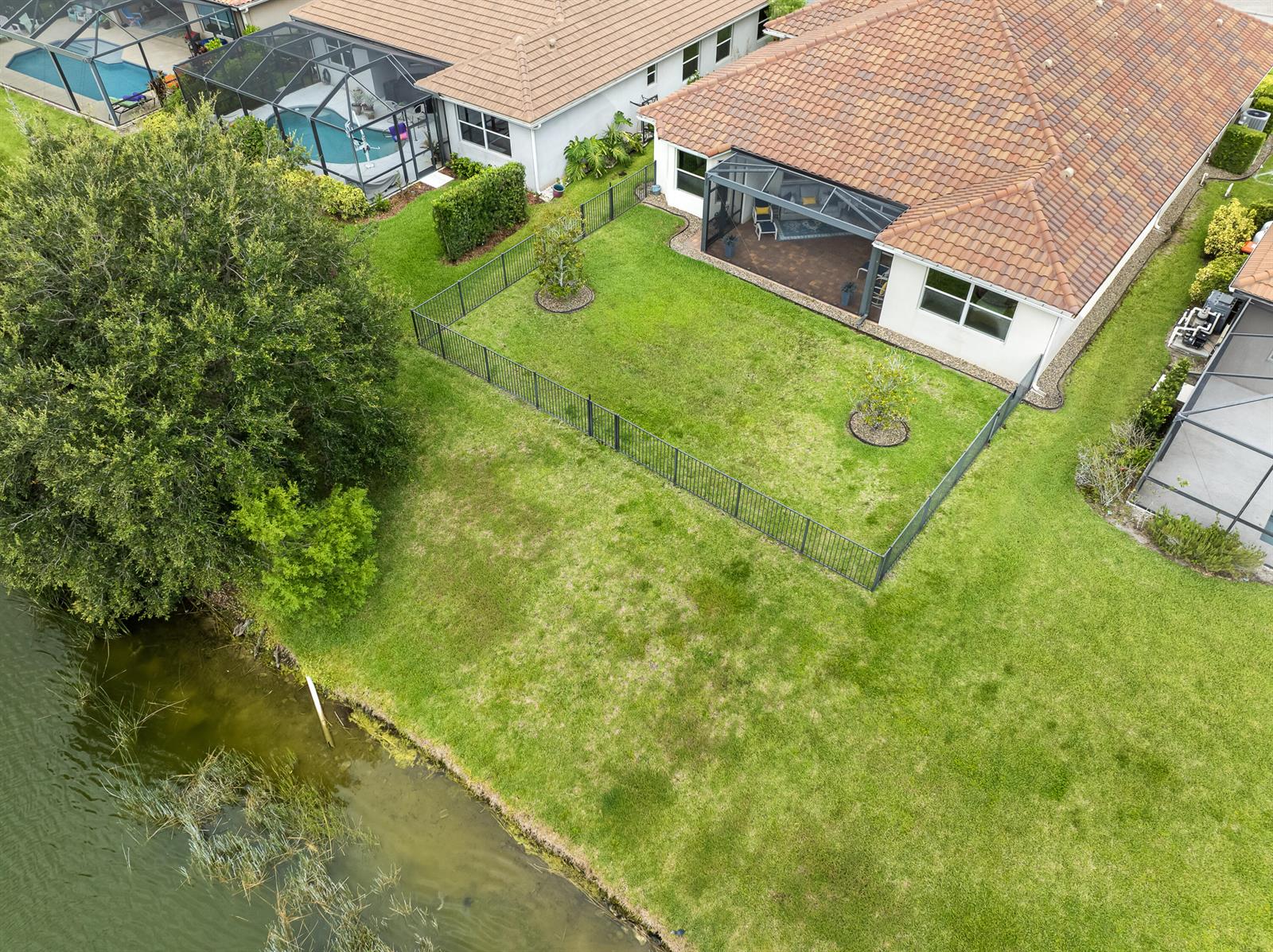 An aerial view shows a fenced backyard with a patio and lawn bordering a lake — 1774 Belmont Circle SW, Vero Beach FL 32968, VERO BEACH, FL