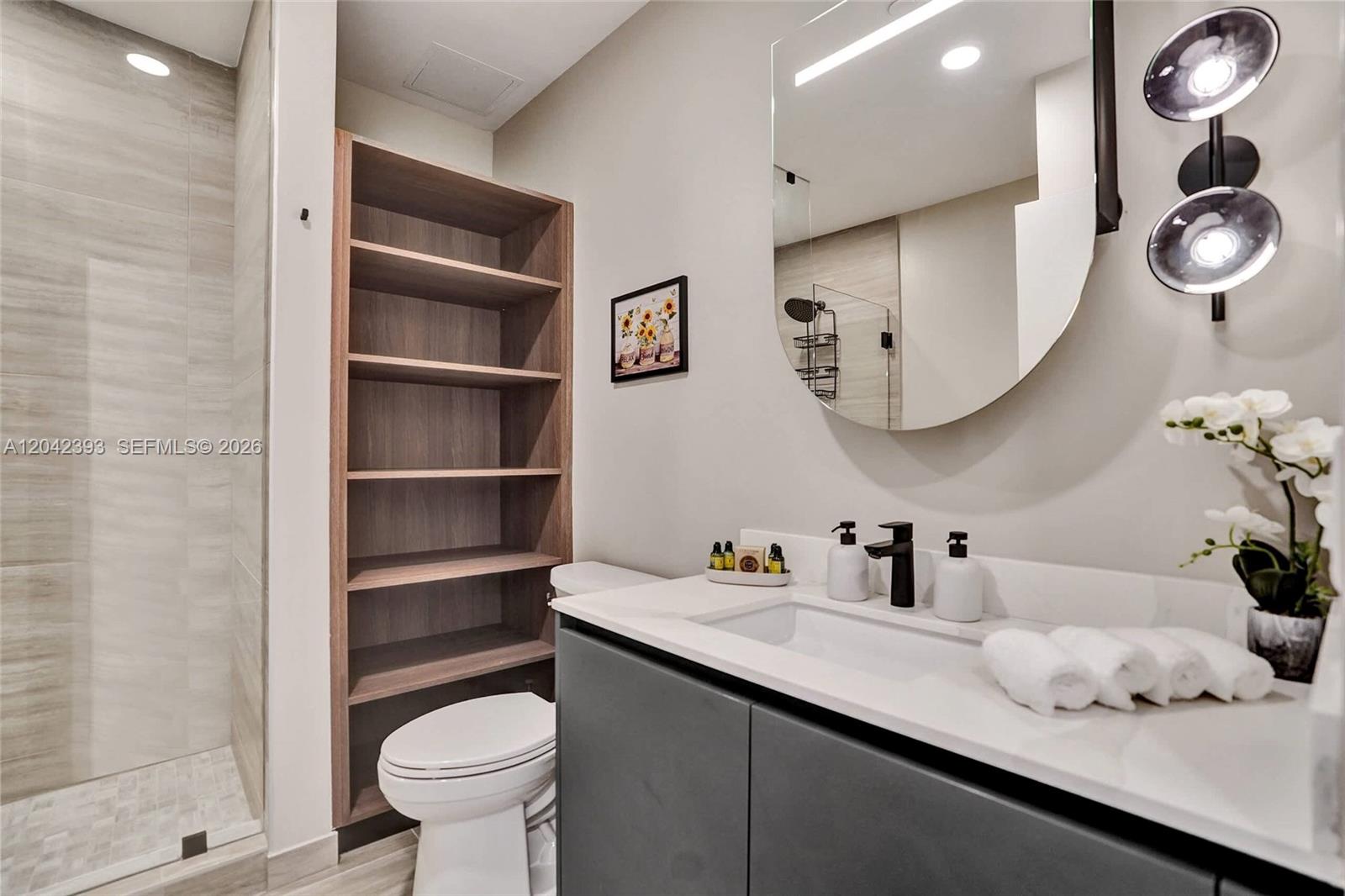 A modern bathroom features a gray vanity, open wood shelving, and a round mirror — 580 72nd St # 1811, Miami Beach FL 33141, MIAMI BEACH, FL