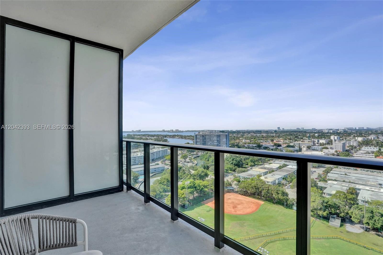 A high-rise balcony overlooks a baseball field, urban landscape, and a distant body of water — 580 72nd St # 1811, Miami Beach FL 33141, MIAMI BEACH, FL