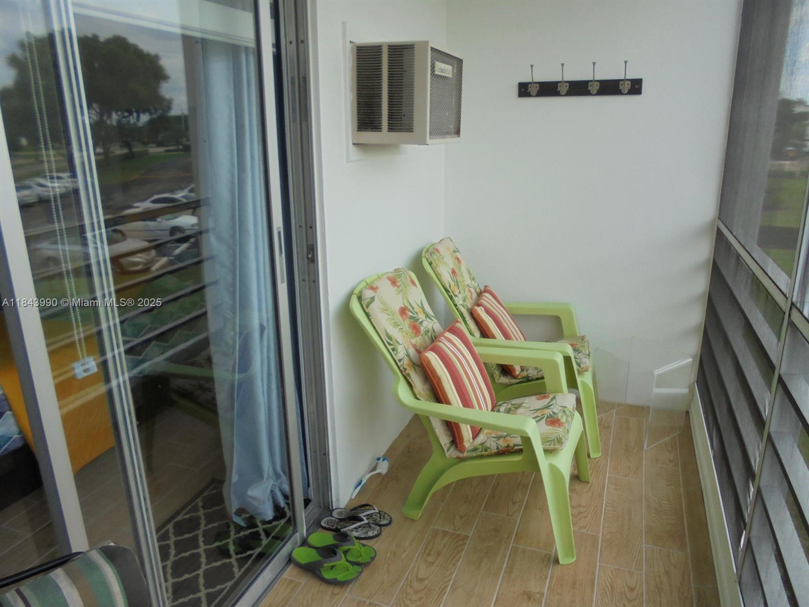 A balcony features two green chairs with floral cushions, a wall-mounted air conditioner, and flooring — 400 Brighton J # 400, Boca Raton FL 33434, BOCA RATON, FL