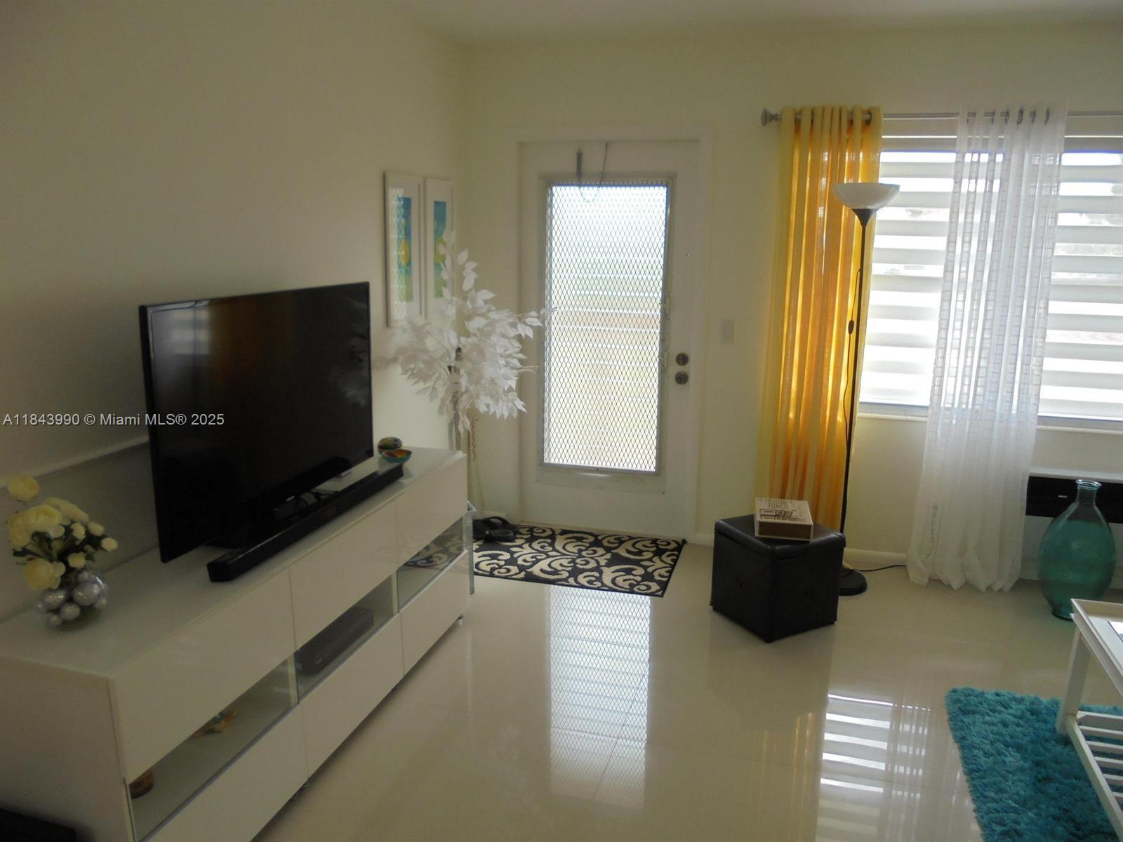 A living room features a white media console, a television, a door, and tile flooring — 400 Brighton J # 400, Boca Raton FL 33434, BOCA RATON, FL