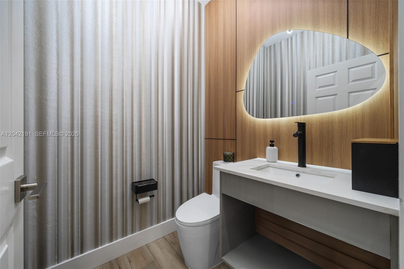 A modern powder room features wood-paneled walls, a backlit organic-shaped mirror, and a floating vanity — 10743 NW 78th Ter, Doral FL 33178, DORAL, FL