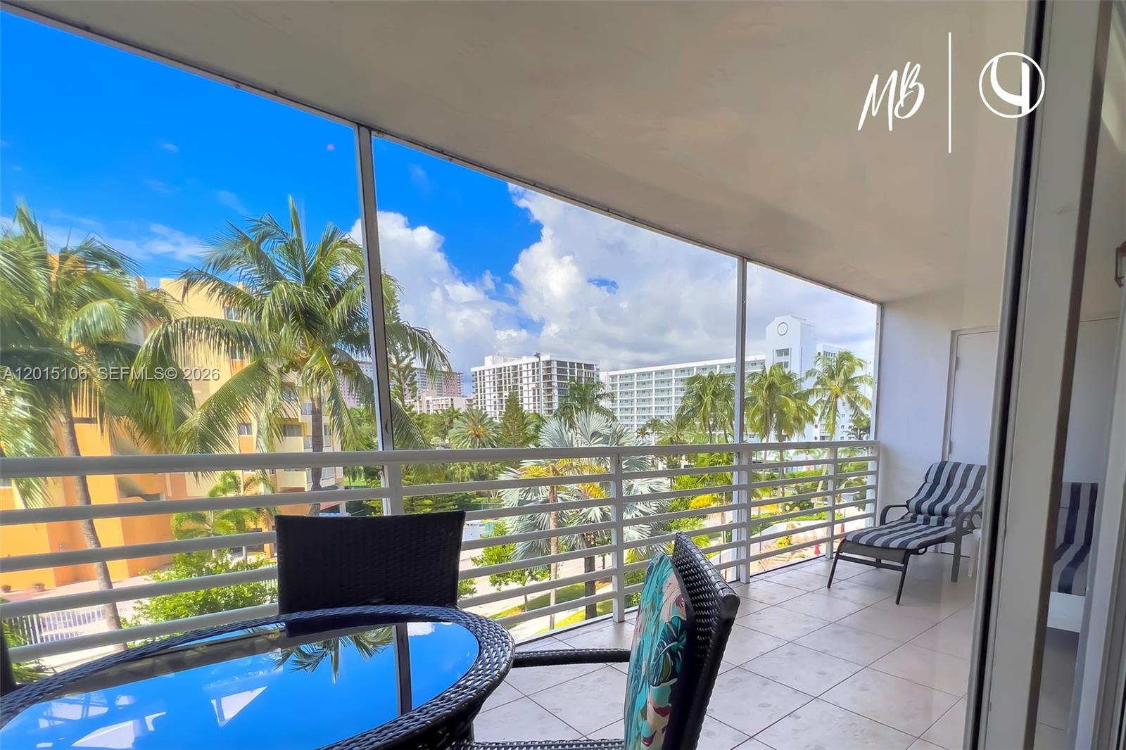 A screened balcony features a glass-topped table, chairs, and a lounge chair overlooking palm trees — SUNNY ISLES BEACH, FL
