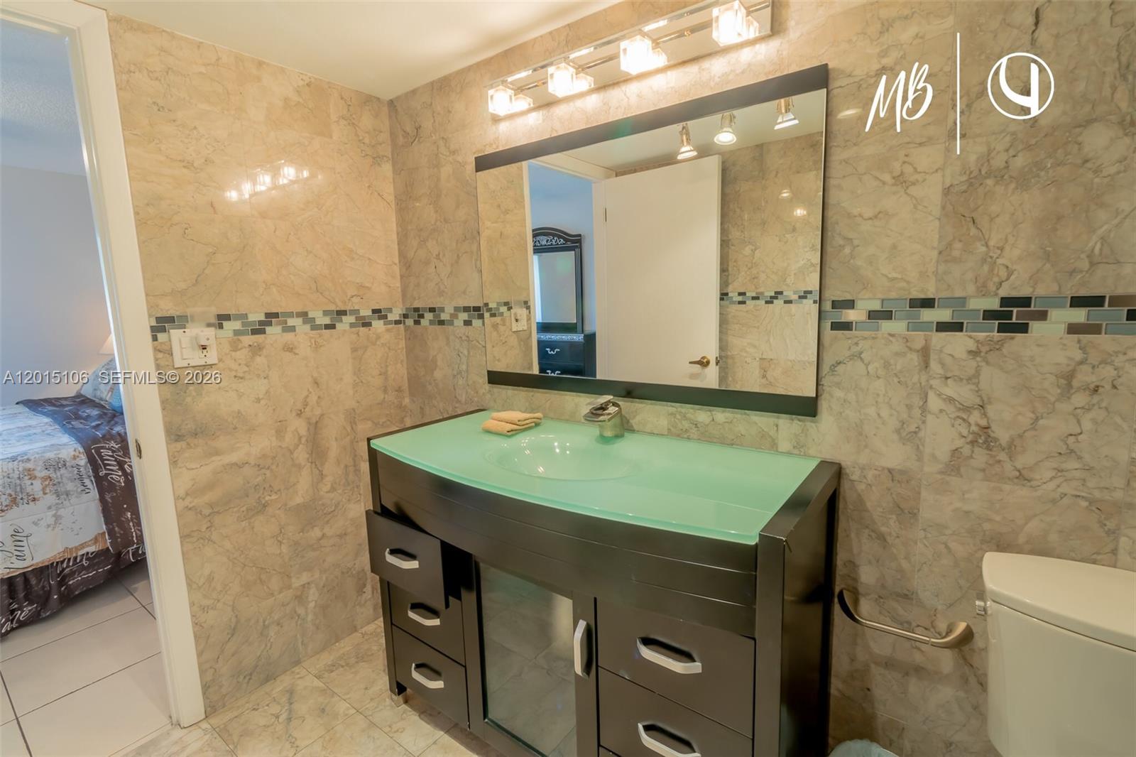 A bathroom features a dark wood vanity with a green glass countertop and marble walls — SUNNY ISLES BEACH, FL