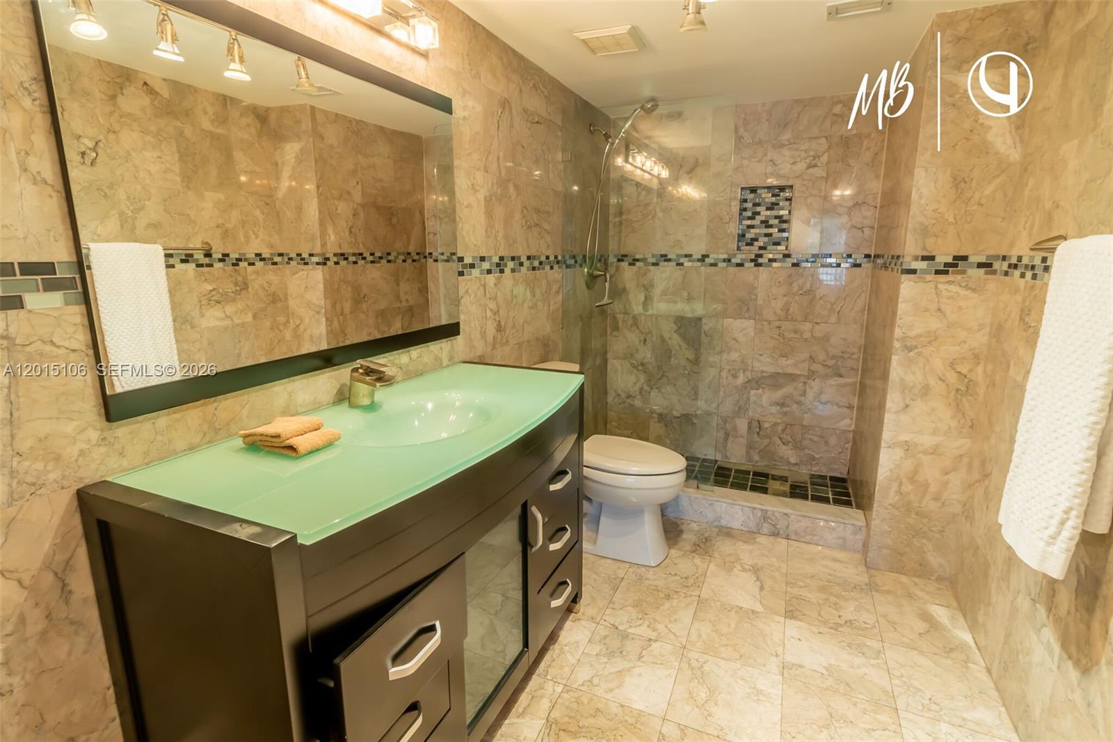 A bathroom features a glass vanity top, dark wood cabinetry, and beige marble wall tiles — SUNNY ISLES BEACH, FL