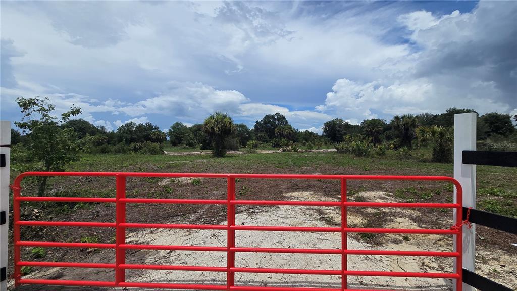 A red metal gate opens to a cleared, grassy lot with scattered trees under a cloudy sky — 21177 NW 268TH STREET, OKEECHOBEE, FL