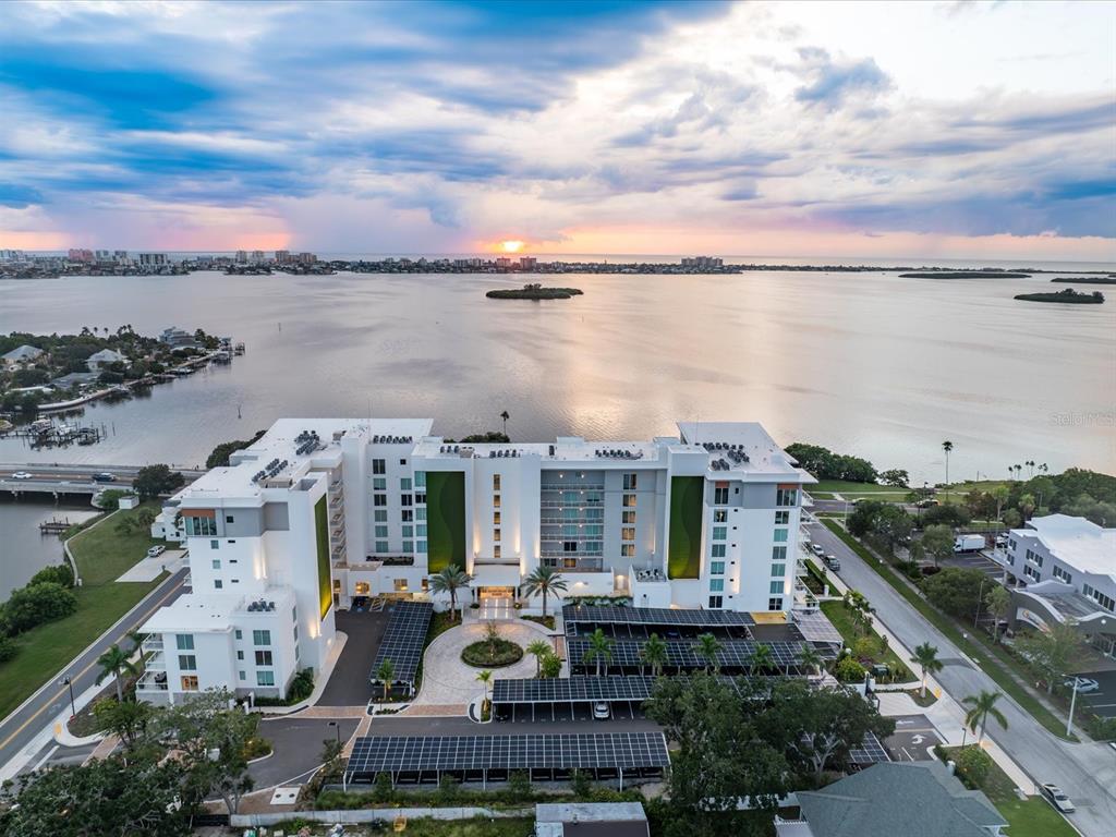 An aerial view shows a modern white apartment building with solar-covered parking lots near water — 1020 SUNSET POINT ROAD UNIT 511, CLEARWATER, FL