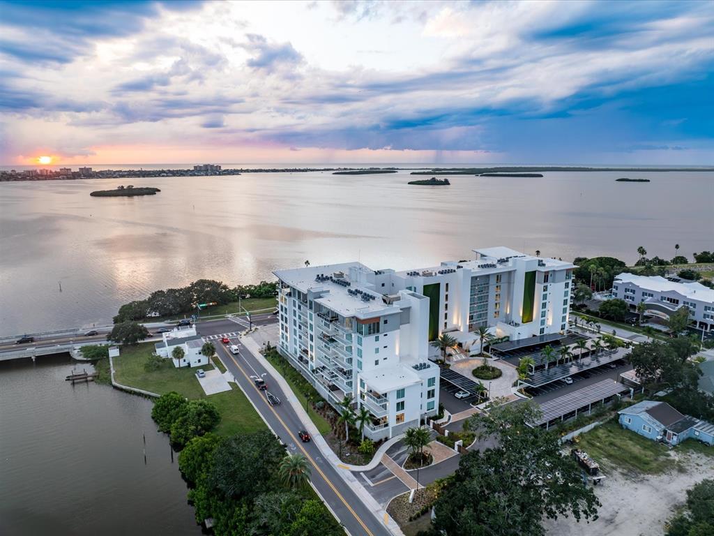 An aerial view shows a modern white apartment building situated along a waterfront at sunset — 1020 SUNSET POINT ROAD UNIT 511, CLEARWATER, FL