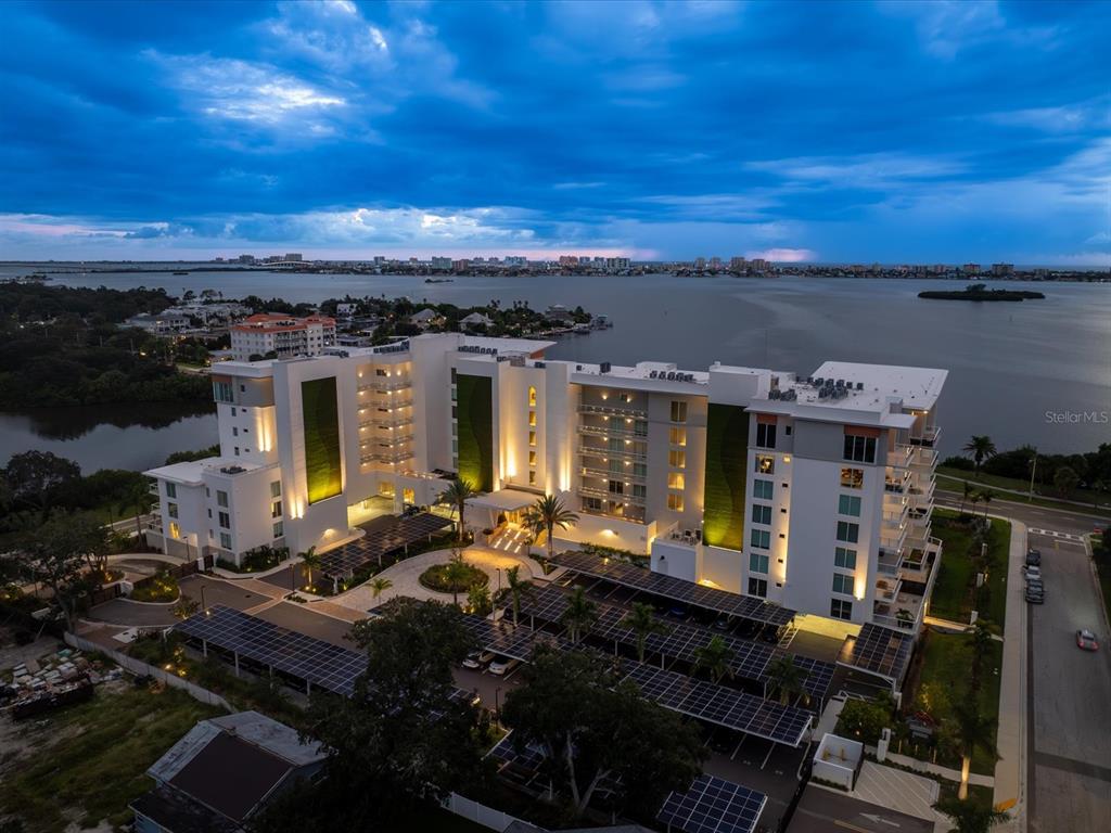 An aerial view shows a modern multi-story apartment building with solar-covered parking at twilight — 1020 SUNSET POINT ROAD UNIT 511, CLEARWATER, FL