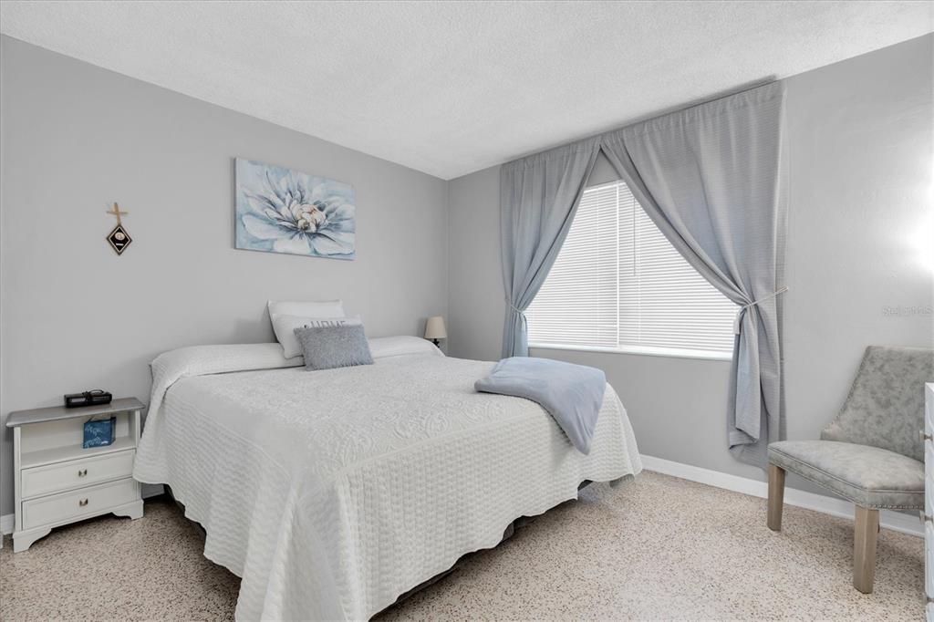 A bedroom features a bed with a white quilt, gray curtains, and a side table — 121 46TH AVENUE UNIT 2A, ST PETE BEACH, FL