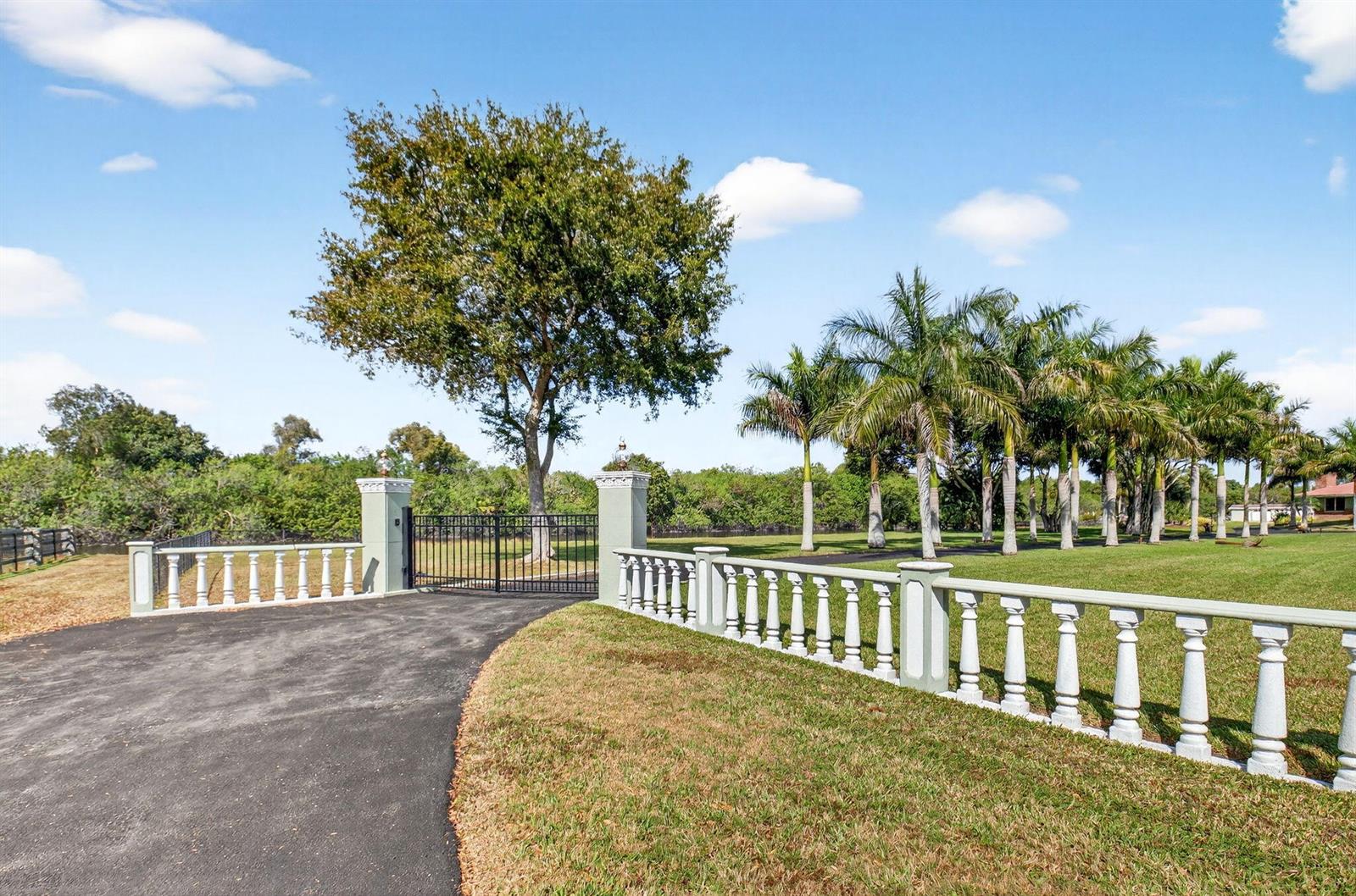 A paved driveway leads to a gated entrance with a white balustrade fence and palm trees — 10965 La Reina Road, Delray Beach FL 33446, DELRAY BEACH, FL