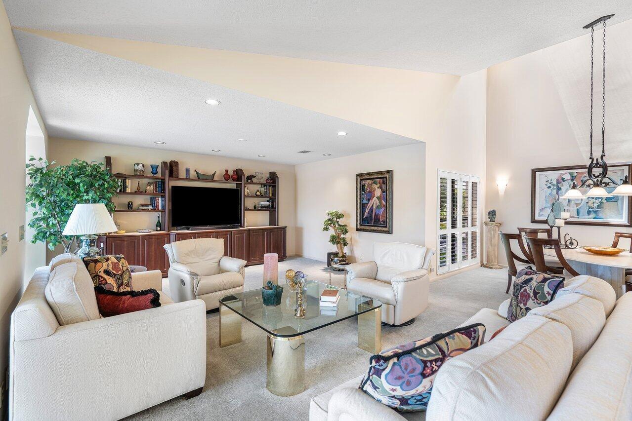 A living room with a vaulted ceiling, beige carpet, white sofas, and a built-in media center — 69 Eastgate Drive Unit D, Boynton Beach FL 33436, BOYNTON BEACH, FL