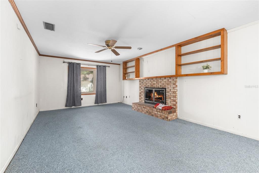 A living room features blue carpeting, white walls, a brick fireplace, and wooden wall shelving — 10503 NW 25TH PLACE, GAINESVILLE, FL