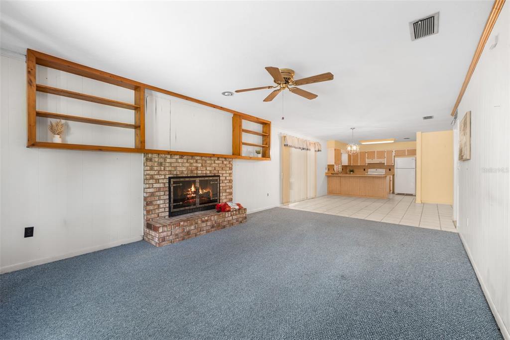 A living room with blue carpet, a brick fireplace, built-in shelving, and an adjacent kitchen — 10503 NW 25TH PLACE, GAINESVILLE, FL