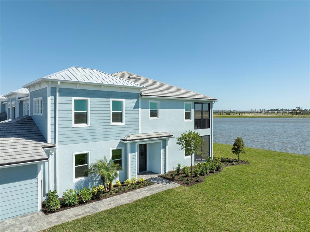 An exterior view of a two-story blue house with a metal roof beside a lake — 12829 JADE EMPRESS LOOP UNIT 101, VENICE, FL