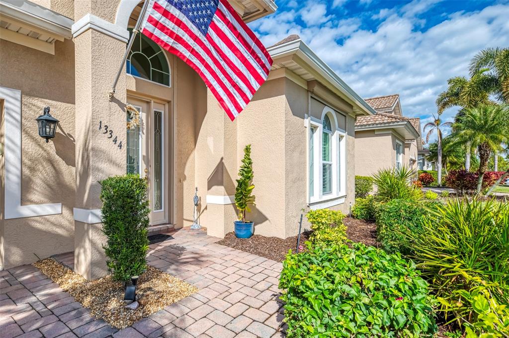 A tan stucco house exterior features a brick walkway, an American flag, and lush landscaping — 13344 CREEKSIDE LANE, PORT CHARLOTTE, FL