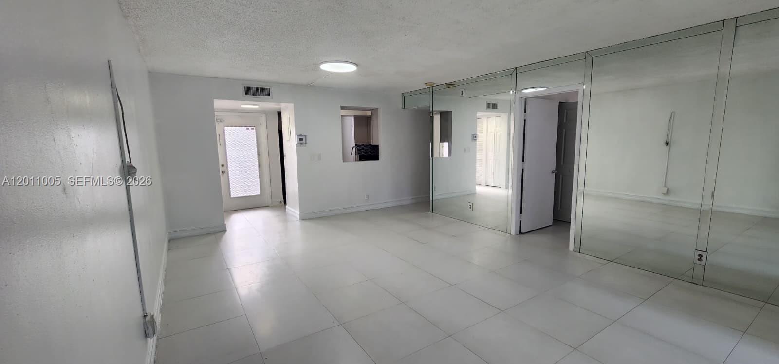 A bright, empty room features white tiled floors, mirrored walls, and a pass-through kitchen window — 1020 NW 80th Ave # 203, Margate FL 33063, MARGATE, FL