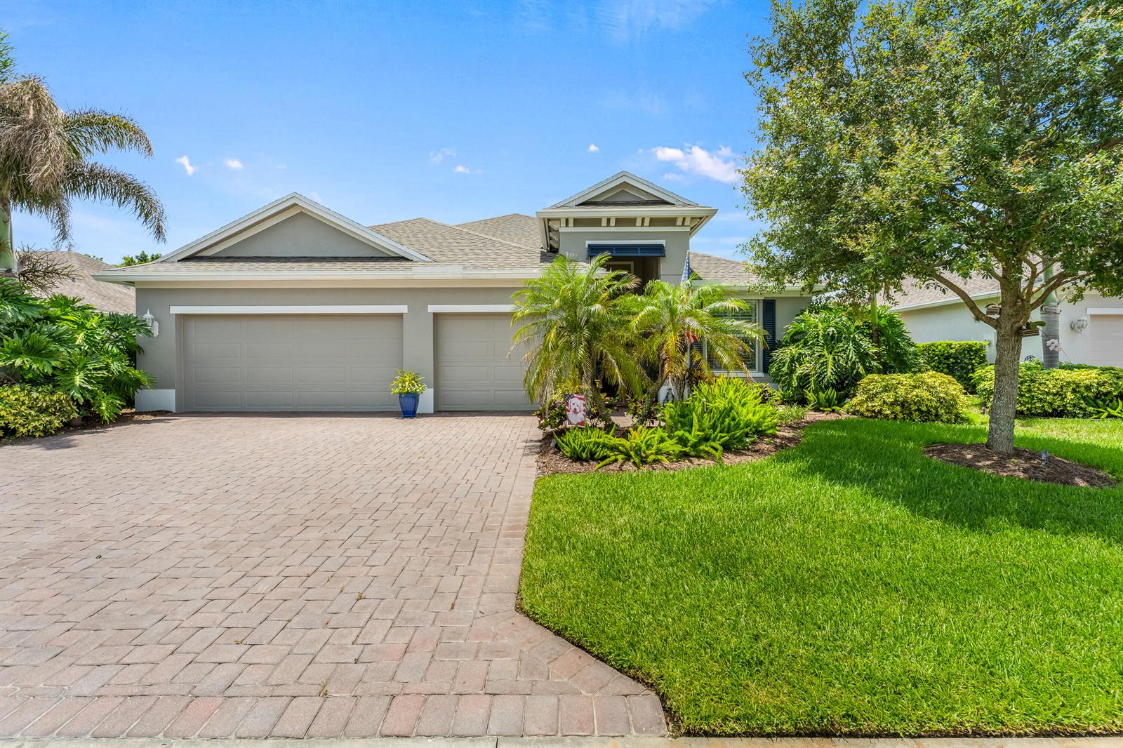 A single-story house with a paved driveway, three-car garage, and lush landscaping under a blue sky — 1573 Segovia Circle, Vero Beach FL 32966, VERO BEACH, FL